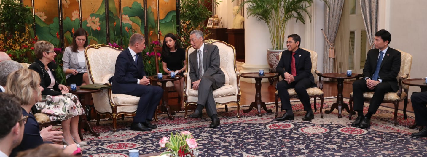 Lee Hsien Loong and others seated in ornate chairs converse in a room with a large floral rug.